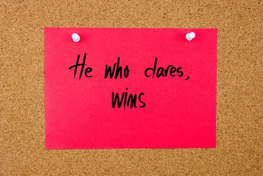 Red paper note with handwritten text He Who Dares Wins pinned on cork board with white thumbtacks