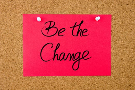 Red paper note with handwritten text Be The Change pinned on cork board with white thumbtacks