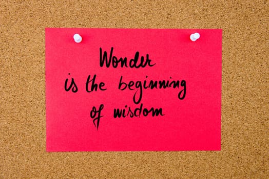 Red paper note with handwritten text Wonder Is The Beginning Of Wisdom pinned on cork board with white thumbtacks