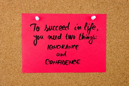Red paper note with handwritten text To Succeed In Life You Need Two Things IGNORANCE and CONFIDENCE pinned on cork board with white thumbtacks