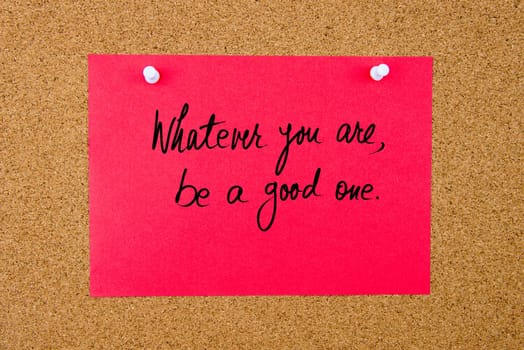 Red paper note with handwritten text Whatever You Are, Be A Good One pinned on cork board with white thumbtacks