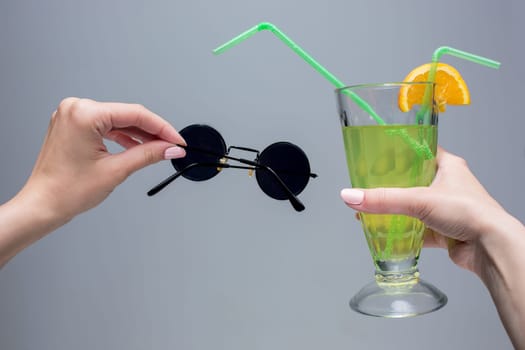 The female hands with cocktail and sunglasses on a gray background