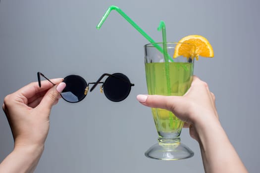 The female hands with cocktail and sunglasses on a gray background