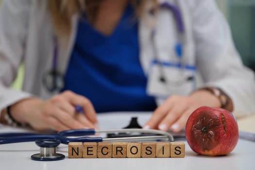 A medical professional conducts an examination of necrosis, symbolically represented by an apple and wooden letters