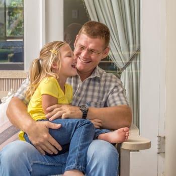 A father sits on a bench on the porch of his house while having fun with his young daughter who is sitting in his lap.