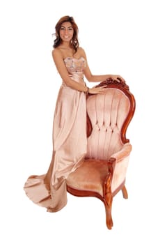 A lovely young woman in a gold color evening dress standing on a pink .armchair, isolated for white background..
