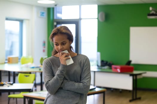 Teenage girl standing in an empty classroom with a mobile phone in her hand. She is looking sad with her arms folded.