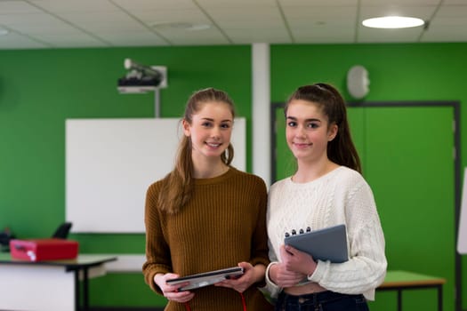 Two teenage girls standing together in a classroom with school eqipment in their hands.