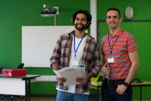 Two male teachers standing together with paper work and a digital tablet. They are smiling and looking at the camera.