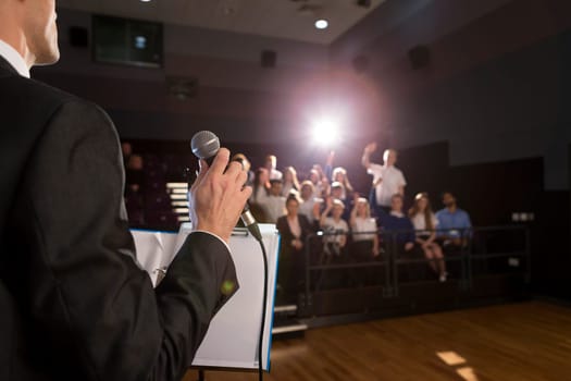 Male teacher making a speech in a lecture hall. He is standing at the podium and some students have their hand up to ask a question.
