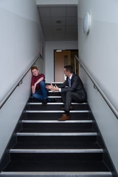 Male student is sitting on the stairs in his school, with his teacher next to him. He is telling him off for his behaviour.