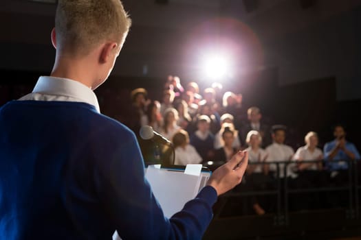 Male student making a speech. He is standing at the podium and is talking to the crowd of people.