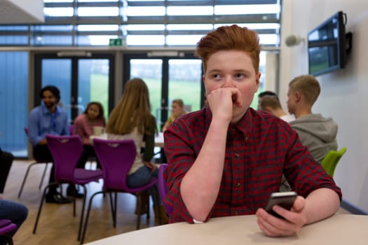 Stressed schoolboy sitting away from people at school. He has a smartphone in his hand and is looking away with his hand to his face.