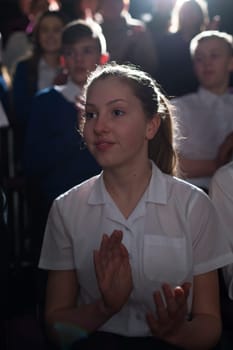Teenage student sitting in a lecture hall in a crowd of students, clapping her hands. She is looking towards the speaker, who can't be seen.