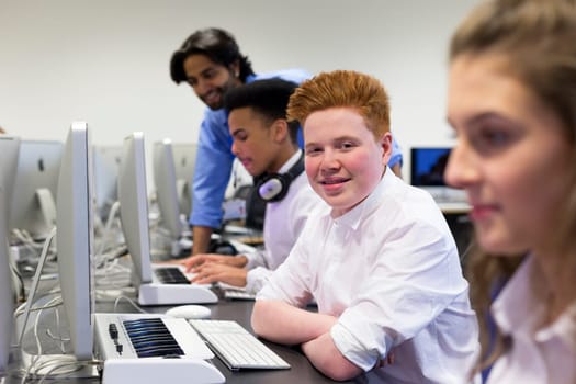 Students using computers in a school lesson. One student is smiling at the camera. There is a teacher helping another student in the background.