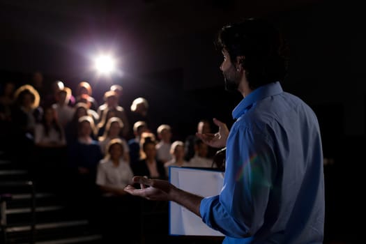 Male teacher giving a speech in a lecture hall to students and teachers.