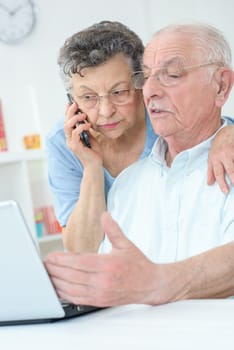 Elderly couple using laptop, wife on telephone
