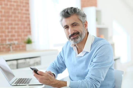 Trendy mature man using smartphone in front of laptop