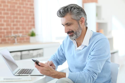 Trendy mature man using smartphone in front of laptop