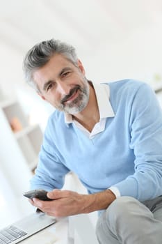 Trendy mature man using smartphone in front of laptop
