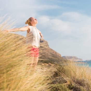 Relaxed woman, arms rised, enjoying sun, freedom and life an a beautiful beach. Young lady feeling free, relaxed and happy. Concept of vacations, freedom, happiness, enjoyment and well being.