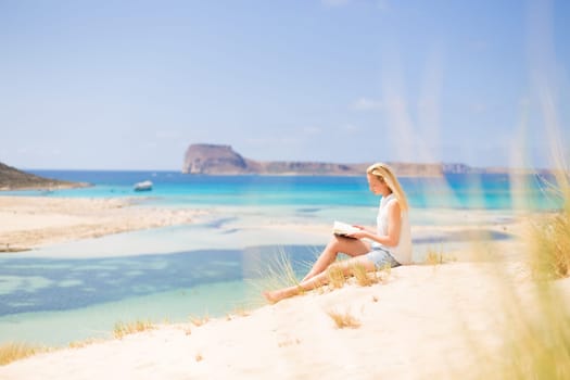 Relaxed woman enjoying sun, freedom and good book an beautiful sandy beach of Balos in Greece. Young lady reading, feeling free and relaxed. Vacations, freedom, happiness, enjoyment and well being.