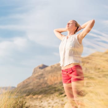 Relaxed woman, arms rised, enjoying sun, freedom and life an a beautiful beach. Young lady feeling free, relaxed and happy. Concept of vacations, freedom, happiness, enjoyment and well being.