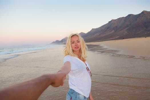 Young romantic couple, holding hands, having fun on perfect deserted beach at sunset. Shot from boyfrieds perspective. Guy looking at her beautiful carefree girlfriend.