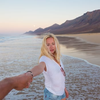 Young romantic couple, holding hands, having fun on perfect deserted beach at sunset. Shot from boyfrieds perspective. Guy looking at her beautiful carefree girlfriend.