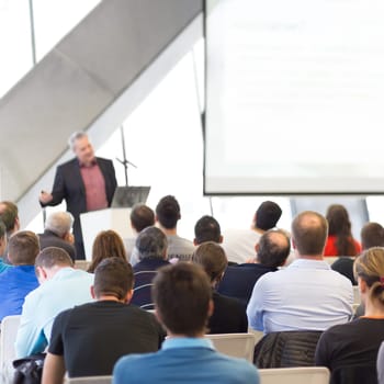 Man giving presentation in lecture hall. Male speeker having talk at public event. Participants listening to lecture. Rear view, focus on people in audience.