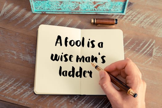 Retro effect and toned image of a woman hand writing on a notebook. Handwritten quote A fool is a wise man&#39 s ladder as inspirational concept image