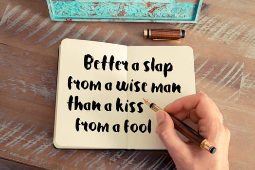 Retro effect and toned image of a woman hand writing on a notebook. Handwritten quote Better a slap from a wise man than a kiss from a fool as inspirational concept image