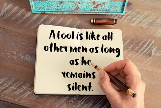 Retro effect and toned image of a woman hand writing on a notebook. Handwritten quote A fool is like all other men as long as he remains silent as inspirational concept image