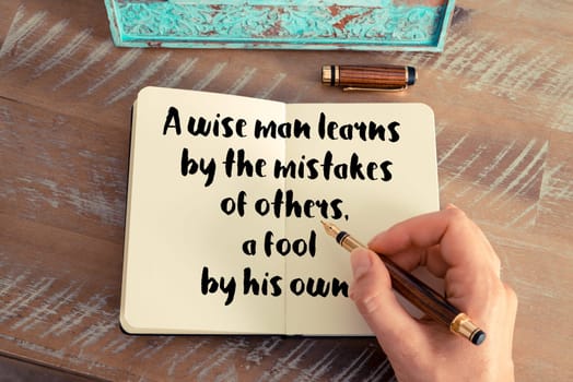 Retro effect and toned image of a woman hand writing on a notebook. Handwritten quote A wise man learns by the mistakes of others, a fool by his own as inspirational concept image