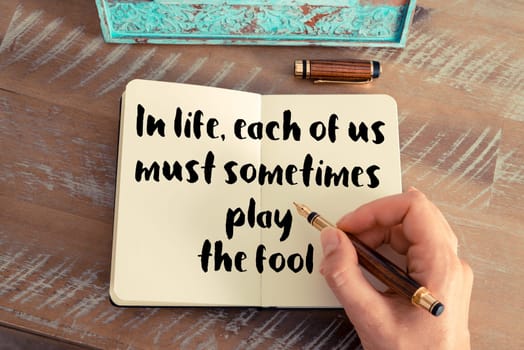 Retro effect and toned image of a woman hand writing on a notebook. Handwritten quote In life, each of us must sometimes play the fool as inspirational concept image