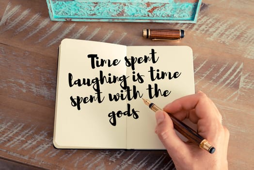 Retro effect and toned image of a woman hand writing on a notebook. Handwritten quote Time spent laughing is time spent with the gods as inspirational concept image