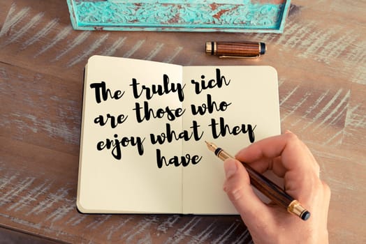 Retro effect and toned image of a woman hand writing on a notebook. Handwritten quote The truly rich are those who enjoy what they have as inspirational concept image