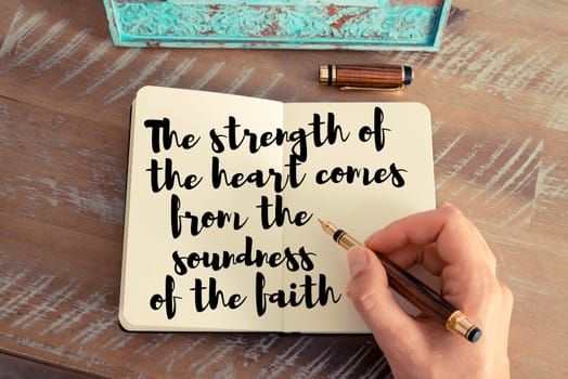 Retro effect and toned image of a woman hand writing on a notebook. Handwritten quote The strength of the heart comes from the soundness of the faith as inspirational concept image