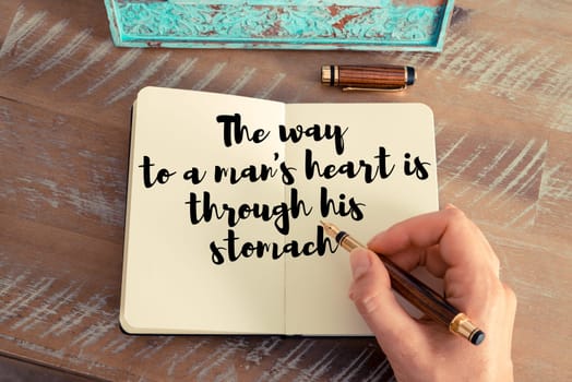 Retro effect and toned image of a woman hand writing on a notebook. Handwritten quote The way to a man&#39 s heart is through his stomach as inspirational concept image