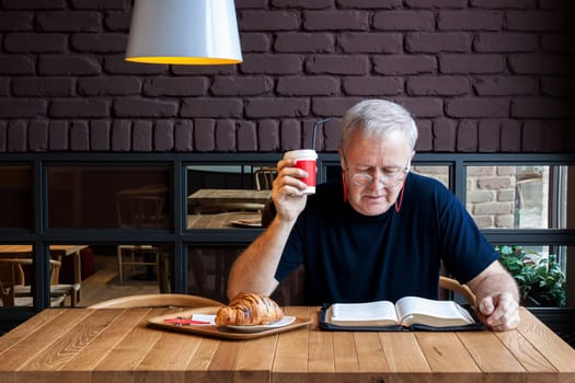 Man sitting at a table with a coffee and a croissant in a snack bar and reading the Bible. Working break dedicated to material and spiritual feeding.