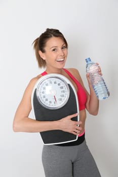Cheerful fitness girl with bottle of water, holding scale, isolated