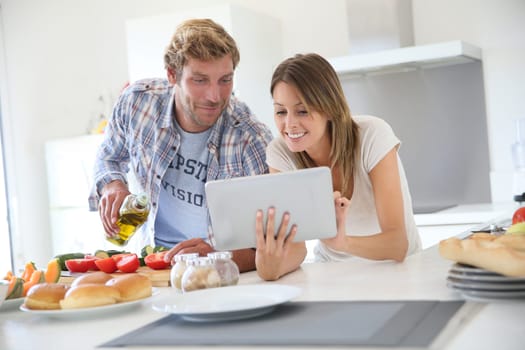 Cheerful couple in kitchen cooking dinner, using tablet