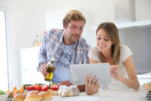 Cheerful couple in kitchen cooking dinner, using tablet