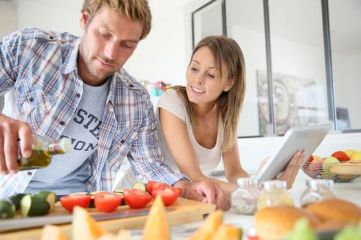 Cheerful couple in kitchen cooking dinner, using tablet