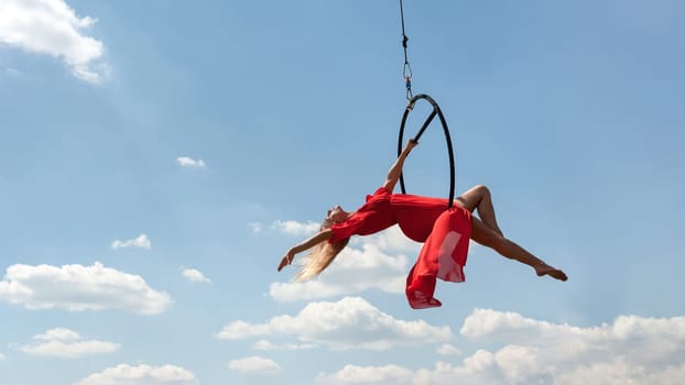 Elegant girl in a red acrobat beautiful dress, performing gymnastic stunts on hoop outdoors against the blue sky with clouds