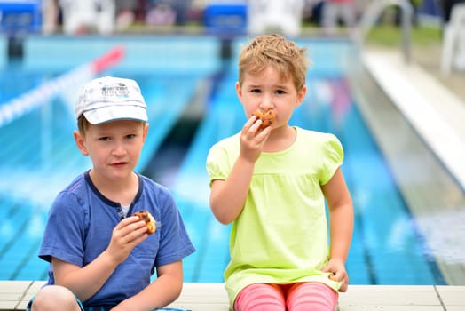 Boy and girl sit at the edge of the pool and eat a muffin
