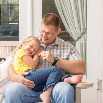 A father and his daughter spend quality time together as they sit on the patio with the father on the bench and his young daughter in his lap.