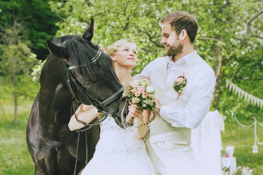 Bride and groom stand together with a magnificent black horse on a summer meadow and rays in the camera, the bride wearing her white dress and holding a bridal bouquet, the groom wearing a beige vest.