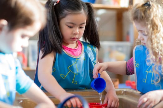 Young children wearing aprons are playing with a water table together in nursery.