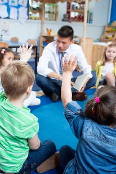 Male teacher giving a lesson to nursery students. They are sitting on the floor in a circle and some have their hands up to ask a question.